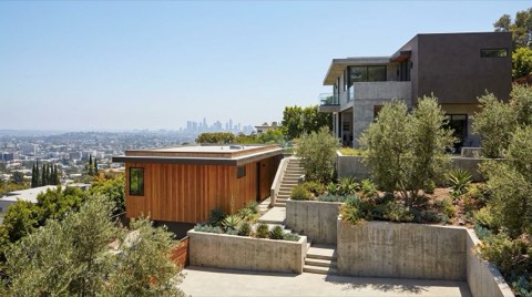 A modern cedar-sided accessory dwelling unit built into a terraced Hollywood Hills hillside next to a larger home, with retaining walls and a hazy city view in daylight