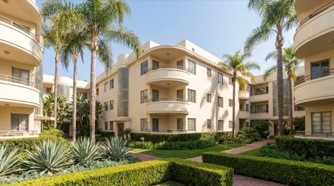 A historic Streamline Moderne courtyard apartment building in Mid-Wilshire with palm trees and a manicured garden under midday sun