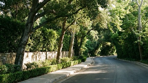 A quiet, shaded sidewalk in Bel Air running alongside stone privacy walls and a curving private road