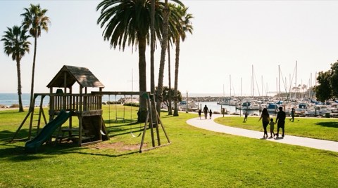 A small coastal neighborhood park with a playground, palm trees, and a path toward the marina