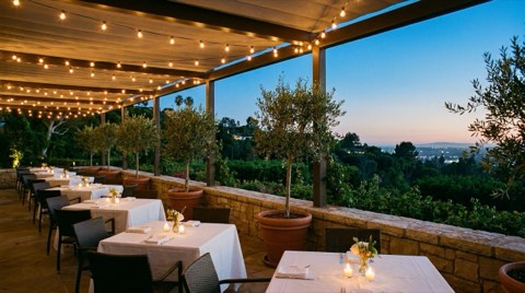 An elegant outdoor restaurant terrace at dusk with string lights, empty candlelit tables, and hillside greenery beyond