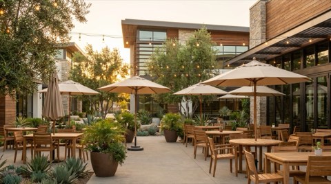 An empty outdoor restaurant patio with string lights, wooden tables, and open umbrellas glowing in warm early-evening light.