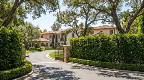 A gated estate driveway with wrought iron gates and tall hedges shielding a large home