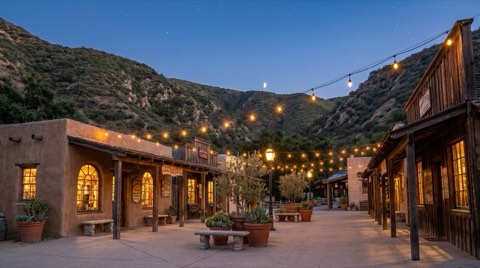 A small canyon village plaza with warm string lights strung between lamp posts and glowing shop windows under a deep blue twilight sky