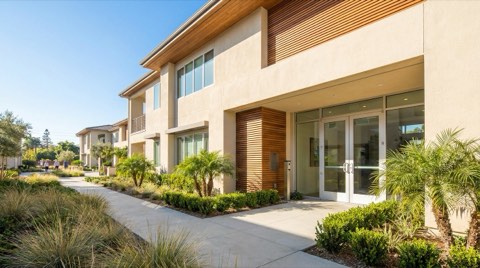 A bright, welcoming modern condo building entrance with glass doors and fresh landscaping framing the walkway in soft morning light.