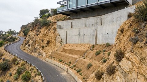 A steep Hollywood Hills canyon street showing a tall engineered retaining wall with visible drainage channels and soil terracing beneath a hillside home's foundation