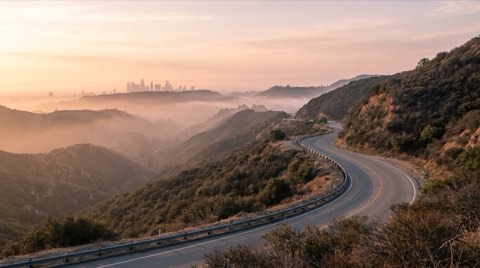 A winding hillside ridge road at dawn with soft pink and gold light, mist in the canyons below, and a hazy Los Angeles skyline visible in the distance