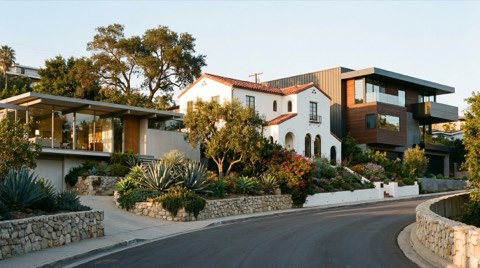 An angled street view in the Hollywood Hills showing a midcentury modern glass home, a white Spanish Colonial Revival home with red tile roof, and a contemporary dark-clad rebuild side by side in late afternoon light