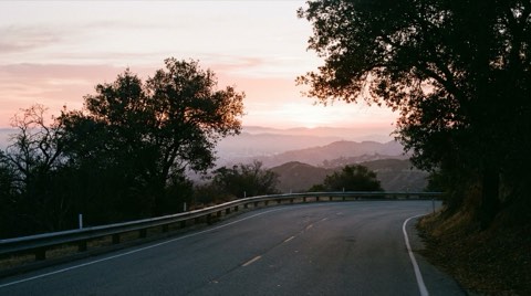 Mulholland Drive winding along a ridge in the Hollywood Hills at dawn, with layered hazy hillsides and the Los Angeles basin below catching soft sunrise light