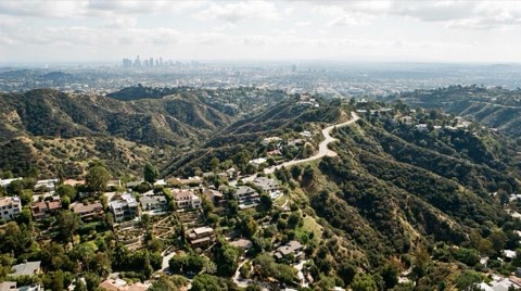 A wide panoramic daytime view of the Hollywood Hills showing layered canyon ridgelines, clusters of hillside homes, and a winding ridge road with the hazy Los Angeles basin beyond
