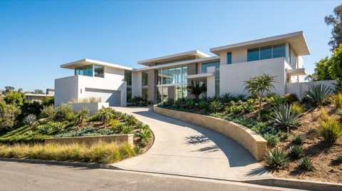 A modern hillside home in the Hollywood Hills with white stucco and glass walls, a curving driveway, and a low retaining wall under clear midday sky