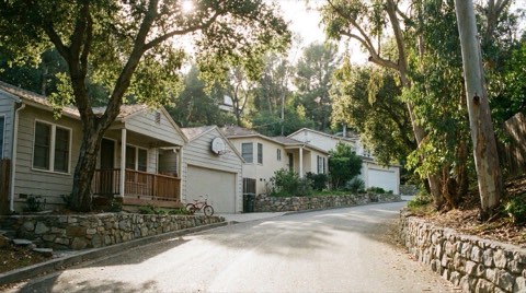 A quiet narrow residential street in the Hollywood Hills on a clear afternoon with modest single-family homes, low stone retaining walls, and a bicycle leaning against a porch railing