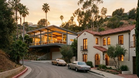 A quiet Hollywood Hills street at golden-hour dusk showing a modern glass-walled home next to an older Spanish-style stucco home with warm lights glowing through the windows