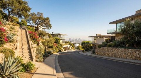 A narrow Hollywood Hills street with a concrete stairway built into the hillside, mature landscaping, and a hazy view of the Los Angeles skyline in the distance