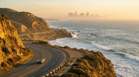 A coastal road along the ocean with the downtown Los Angeles skyline visible hazily in the distance