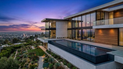 A modern Hollywood Hills luxury home with glass walls and an infinity pool at dusk, with city lights beginning to glow in the valley below