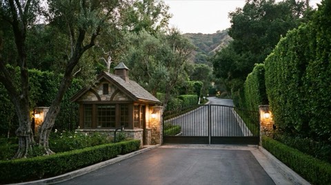 A discreet guardhouse-style gated community entrance at dusk with warm lantern light and dense hedges