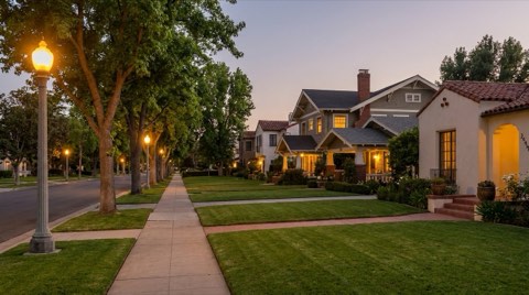 A peaceful residential street in early evening with glowing streetlamps and well-kept sidewalks