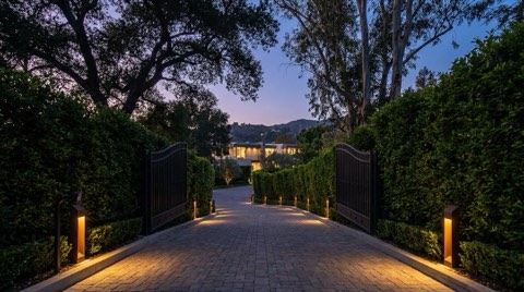 An elegant gated hillside driveway entrance in the Hollywood Hills framed by tall hedges and trees, with warm landscape lighting at dusk