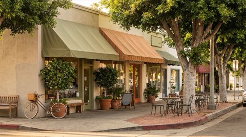 A shaded village street corner in Los Feliz with small boutique storefronts and sidewalk cafe tables in morning light