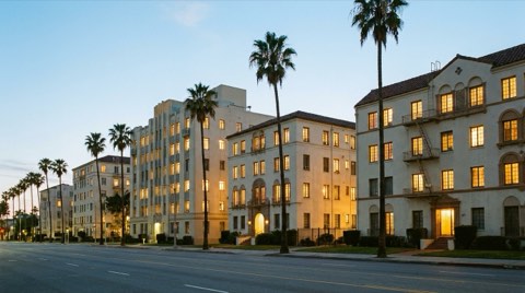 A row of historic mid-rise apartment buildings along a boulevard at dusk with warm glowing windows and palm trees