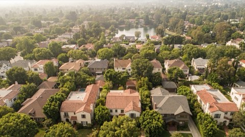 An aerial view of established rooftops among mature trees with a glimpse of a private lake in the distance.