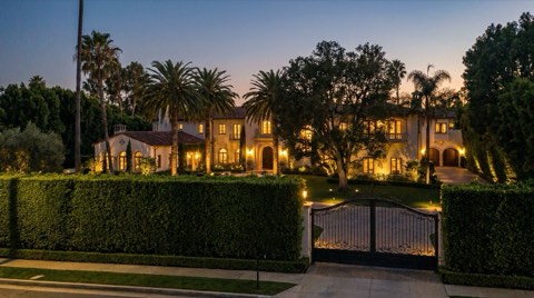 A grand estate-like home seen behind a trimmed hedge and iron gate at dusk.