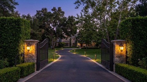 An elegant wrought iron estate gate flanked by manicured hedges with a curving driveway disappearing toward a home obscured by mature trees at dusk
