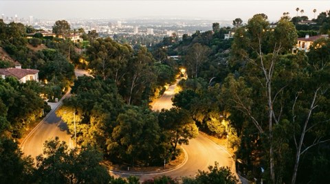 A winding canyon road at dusk descending from a hillside toward distant streetlights