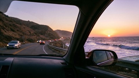 A coastal highway at sunset with ocean waves on one side, evoking a commute along the coast