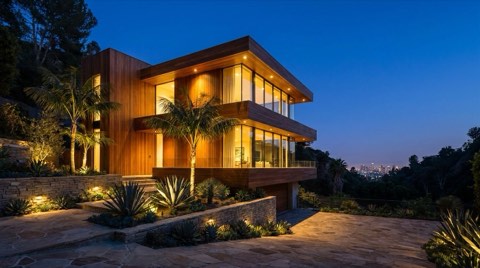 A staged contemporary hillside home glowing with warm interior light at dusk, viewed from a three-quarter angle with landscaped terrace