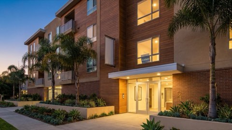 A mid-rise apartment building exterior with a clean entrance and well-kept landscaping glowing at dusk