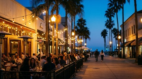 A warmly lit outdoor dining patio on a Santa Monica pedestrian street at dusk with string lights and silhouetted palm trees