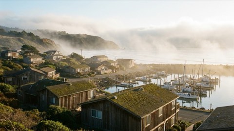 A quiet beach town with low-rise homes between a hillside and the ocean, marina visible in the distance