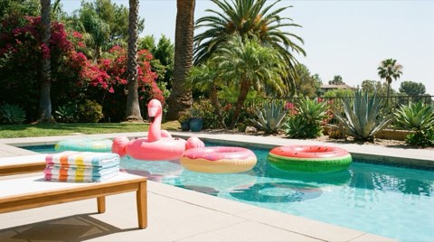 A backyard pool area with colorful pool floats and stacked towels on a lounge chair in bright midday sun