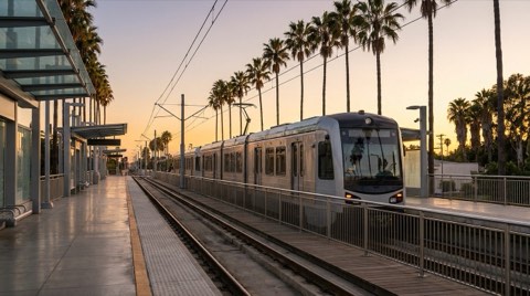 An empty outdoor Expo Line light rail platform in Santa Monica with a train arriving and palm trees along the tracks at late-afternoon light