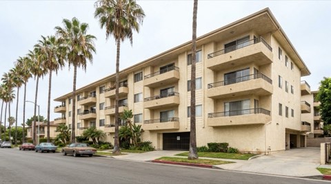 A mid-rise cream stucco apartment building with balconies and palm trees along a Santa Monica street on an overcast day