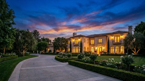 An elegant Bel Air estate exterior at blue hour with warm lights glowing from within and a manicured front garden