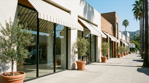 An upscale outdoor boutique retail strip with awnings and potted trees along the sidewalk
