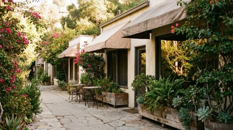 A small boutique canyon-village street with awnings, potted plants, bougainvillea, and outdoor cafe tables in soft afternoon light