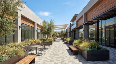 An open-air retail plaza walkway with contemporary storefront architecture, planters, and outdoor seating under bright daylight.