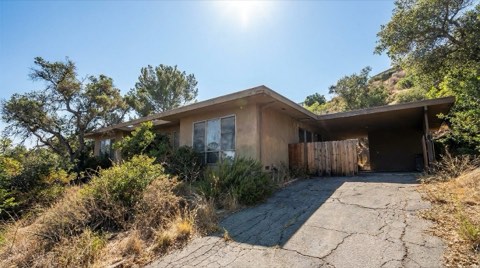 A faded 1960s mid-century hillside home in Hollywood Hills with overgrown landscaping and a cracked driveway, photographed in bright midday sun with renovation potential evident