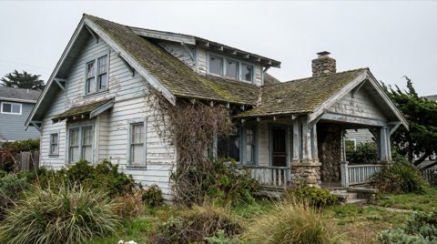 An older beach cottage with peeling paint and an overgrown yard showing renovation potential