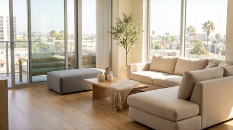 A warmly staged modern living room with neutral furniture, a coffee table, and large windows letting in natural daylight.