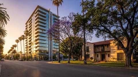 A golden-hour contrast shot of a sleek glass high-rise condo tower beside an older Spanish-style single-family home with a tiled roof and mature trees