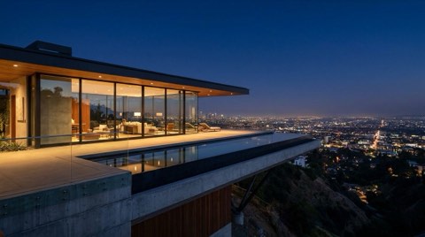 A striking modern glass-walled Hollywood Hills home at night with a cantilevered deck and illuminated infinity pool, overlooking a sweeping carpet of city lights below
