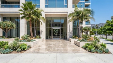 A straight-on daytime view of an upscale condo building entrance with polished stone flooring, a glass door, and palm trees flanking the facade