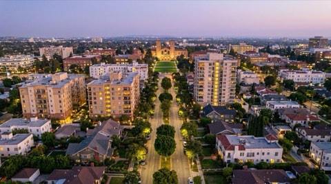 An elevated dusk view down a wide boulevard mixing mid-rise condo towers and tree-lined single-family streets with a university campus in the distance