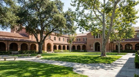 A quiet school campus exterior with a brick building, covered walkways, and a green lawn shaded by mature trees