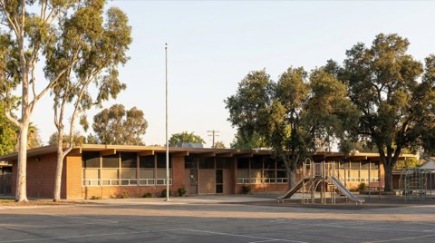 A mid-century public elementary school building with playground equipment under mature trees in morning light.